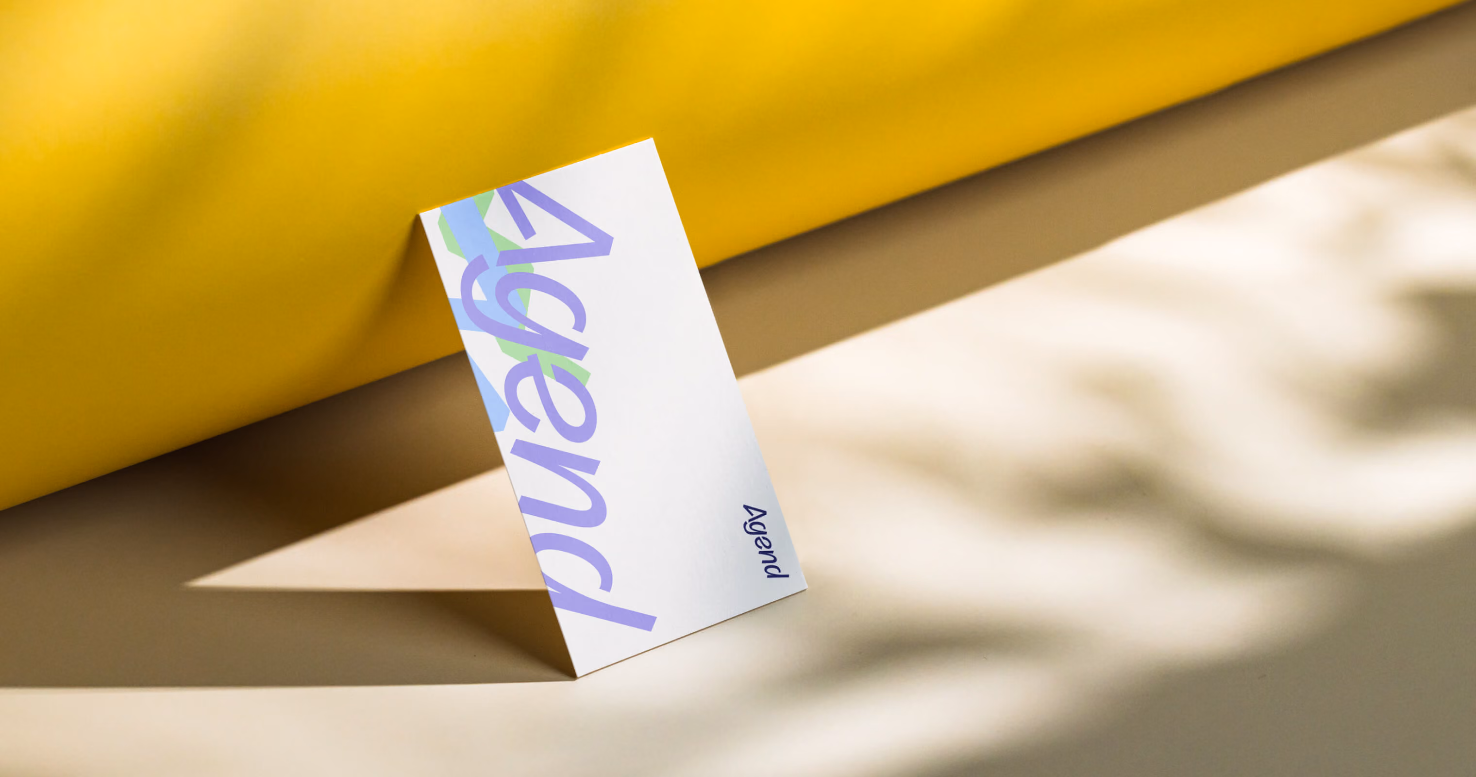 Agend business card with the logotype in lilac and blue, leaning against a yellow surface in soft shadow.