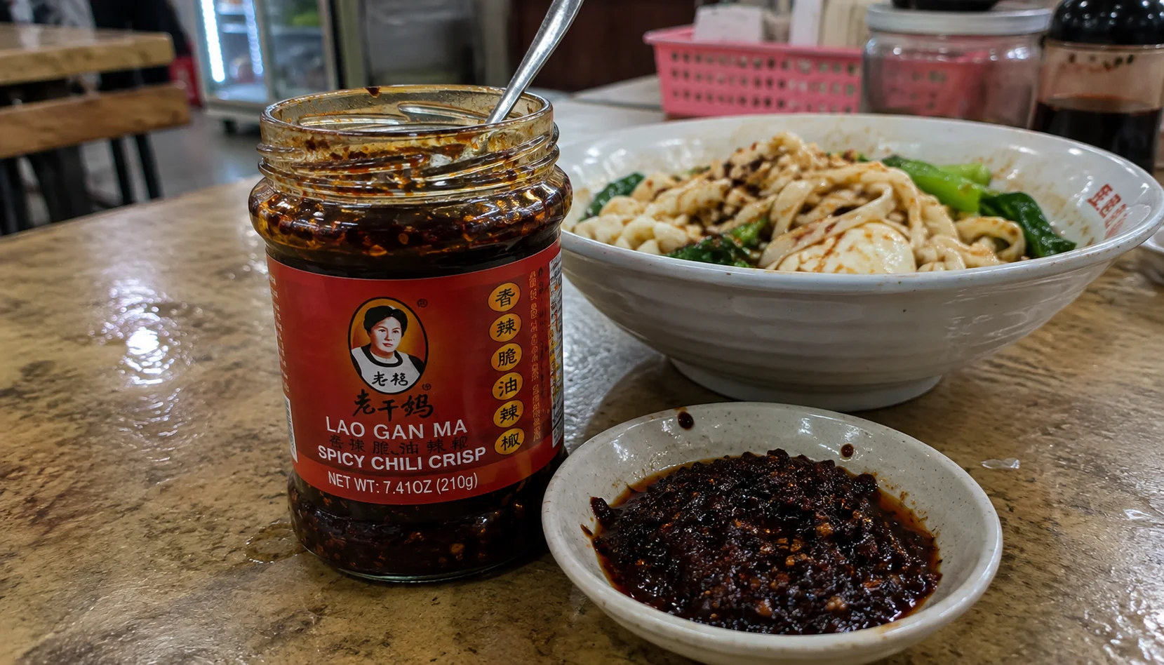 An open jar of Lao Gan Ma spicy chilli crisp on a canteen table, with a saucer of the chilli oil and a bowl of noodles and greens behind