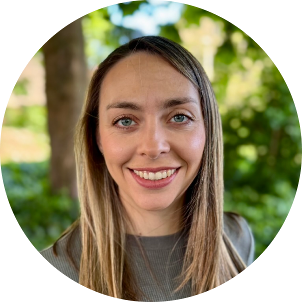 Smiling woman with long blonde hair and blue eyes standing outdoors with green foliage in the background.