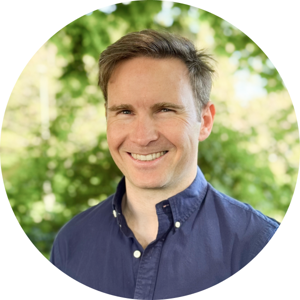 Smiling man with short brown hair wearing a dark blue button-up shirt, standing outdoors with green foliage in the background.