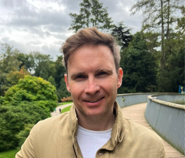 Smiling man with short brown hair wearing a beige jacket and white shirt standing on a curved wooden bridge surrounded by green trees and cloudy sky.