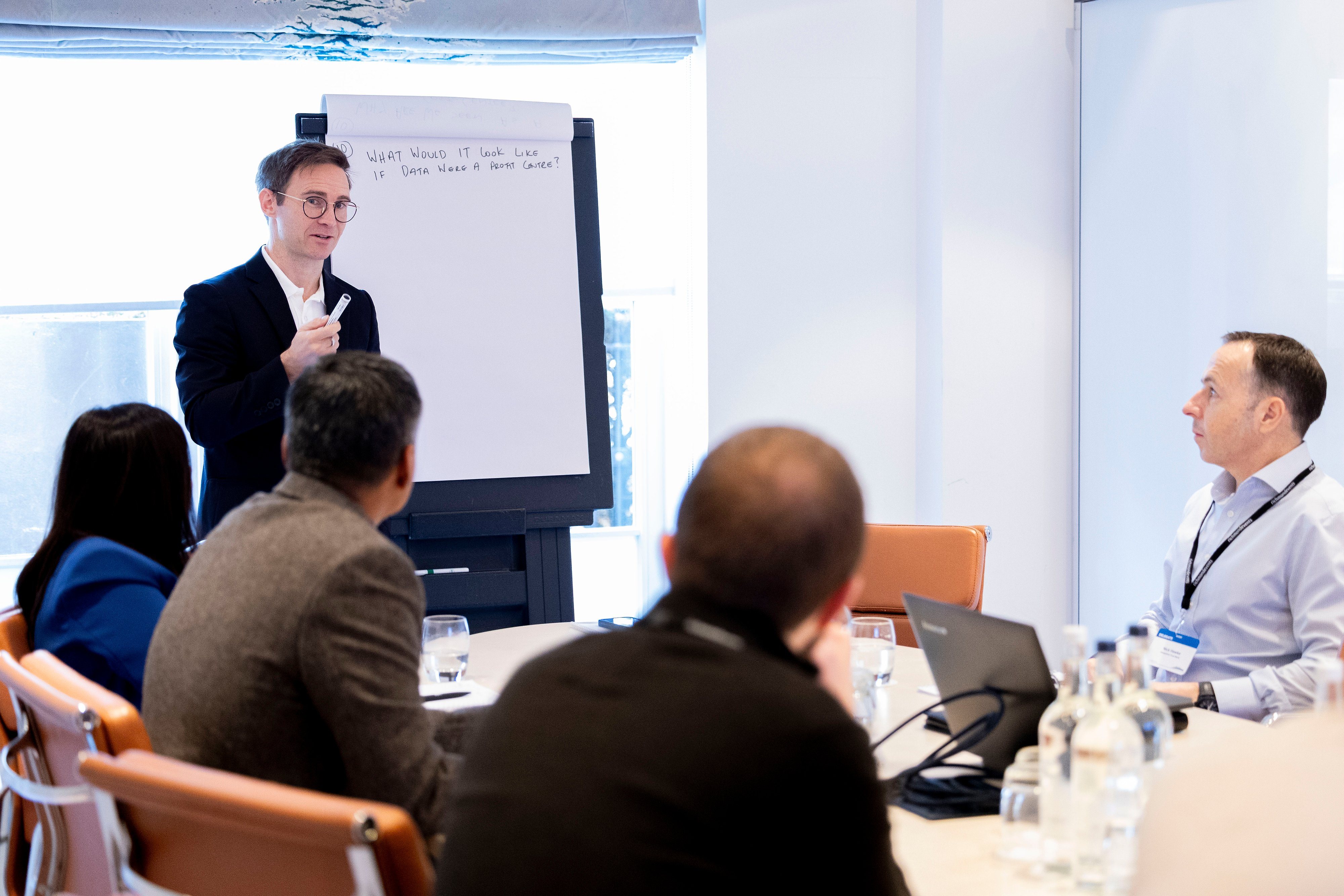 Man in a suit presenting to a group in a conference room, standing next to a flip chart with the question 'What would it look like if data were a profit center?' written on it.