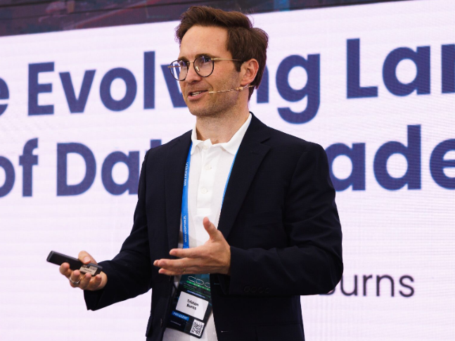 Man wearing glasses and a microphone headset giving a presentation in front of a large screen with partially visible text.