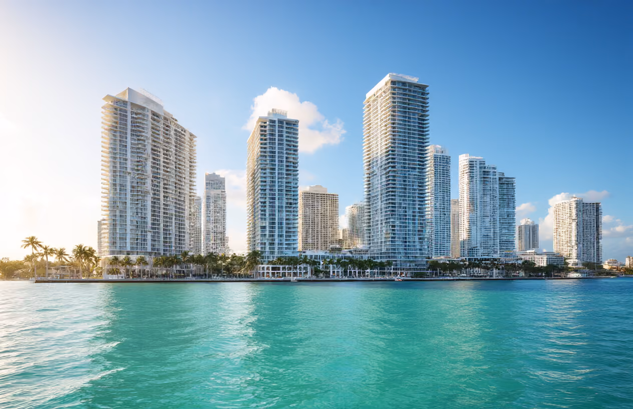 Create a realistic image of Miami's iconic downtown skyline featuring modern luxury high-rise condominiums and hotels along Biscayne Bay, with pristine turquoise waters in the foreground, palm trees lining the waterfront, and a bright sunny day with clear blue skies, showcasing the premium real estate market characteristics of Miami's vacation rental and fractional ownership properties, captured from a water-level perspective that emphasizes the coastal luxury lifestyle and urban sophistication of the area, with warm golden lighting that highlights the glass facades of the buildings reflecting the tropical atmosphere, absolutely NO text should be in the scene.