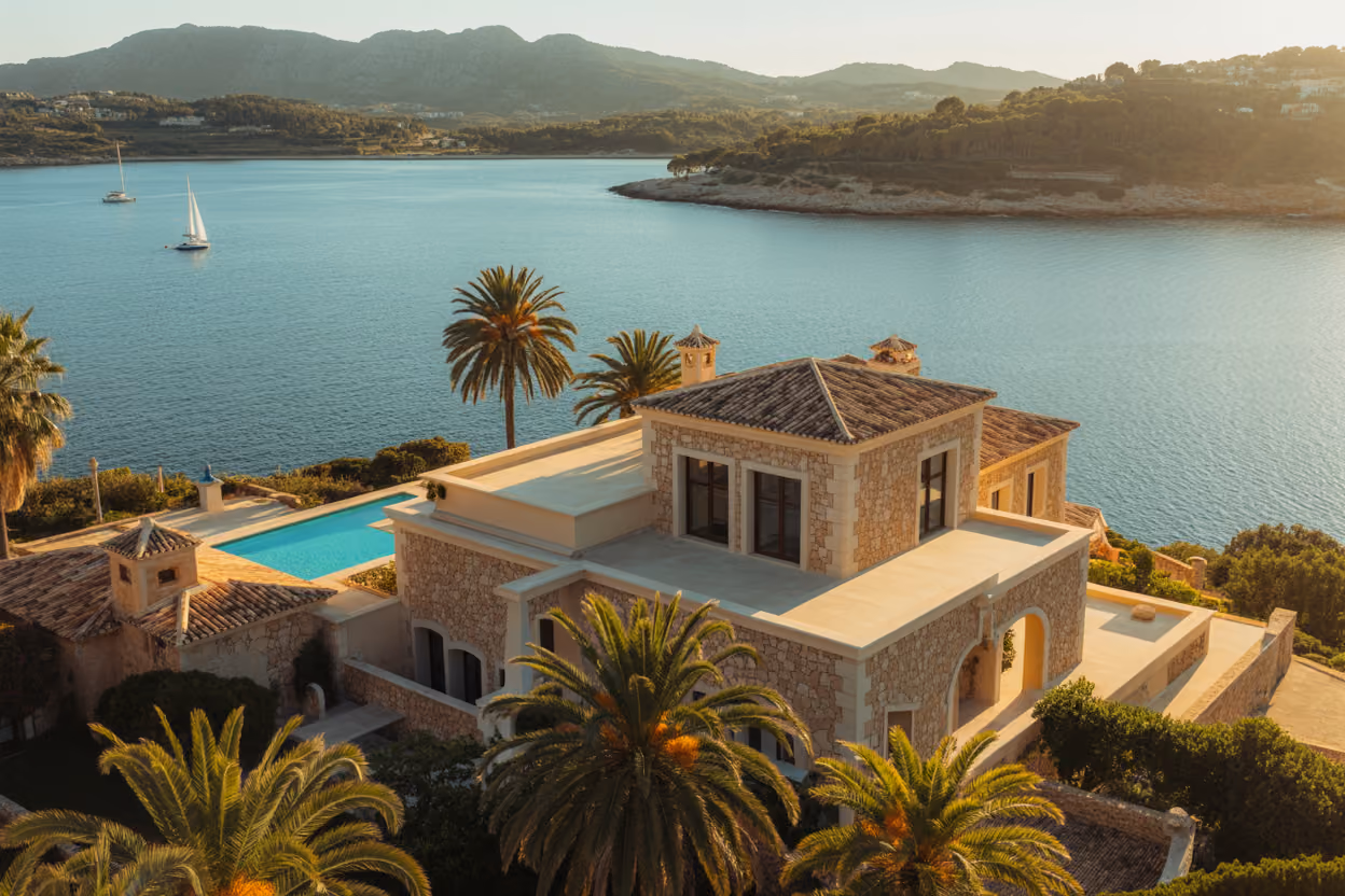 Create a realistic image of a stunning Mediterranean villa with traditional Mallorcan stone architecture overlooking turquoise coastal waters, featuring terracotta roof tiles, white limestone walls, and lush palm trees in the foreground, with golden sunset lighting casting warm glows across the property's infinity pool and expansive terraces, showcasing the luxury and investment potential of Mallorca real estate, captured from an elevated perspective that emphasizes both the property's grandeur and the island's natural beauty, with sailboats visible in the distant bay and rolling hills covered in olive groves extending into the background, creating an aspirational scene that represents successful property investment and Mediterranean lifestyle living, absolutely NO text should be in the scene.