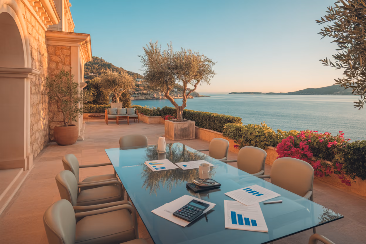 Create a realistic image of a luxurious Mediterranean villa with traditional Mallorcan stone architecture overlooking the turquoise waters of the Balearic Sea, featuring a modern glass conference table on an elegant terrace with financial documents, calculators, and property investment charts spread across it, complemented by potted olive trees and bougainvillea, warm golden hour lighting casting soft shadows, conveying a sophisticated business atmosphere in a premium coastal setting, absolutely NO text should be in the scene.