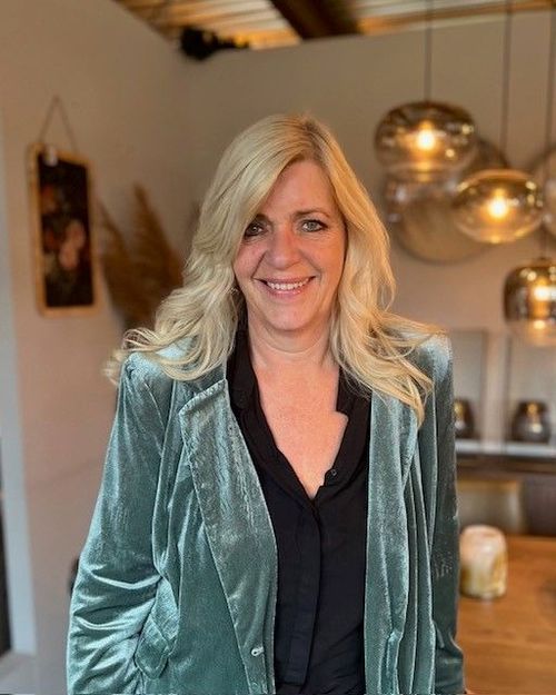 Smiling woman with long blonde hair wearing a green velvet blazer and black blouse standing indoors with warm lighting.