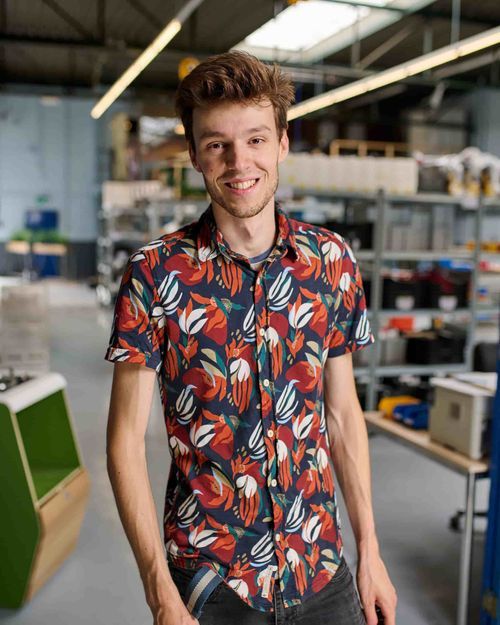 Young man smiling in a colorful floral shirt standing in a workshop or industrial space.