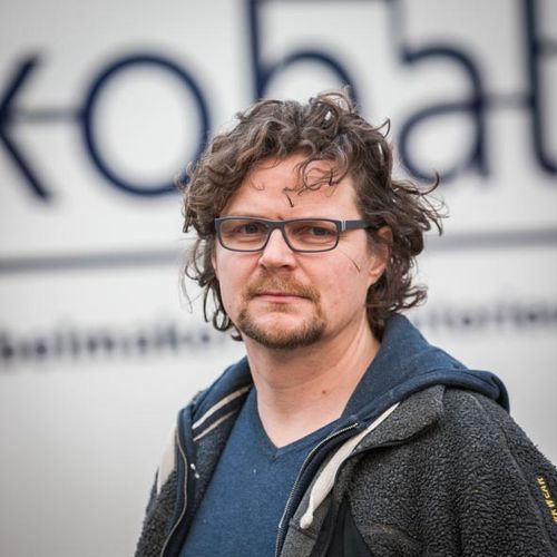 Man with curly hair and glasses wearing a dark jacket and blue shirt standing in front of a blurred sign.