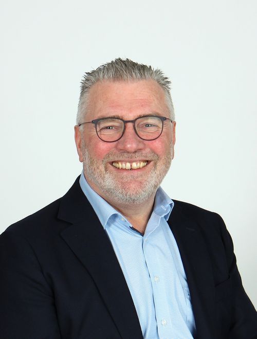 Smiling middle-aged man with gray hair, beard, glasses, wearing a light blue shirt and dark blazer.