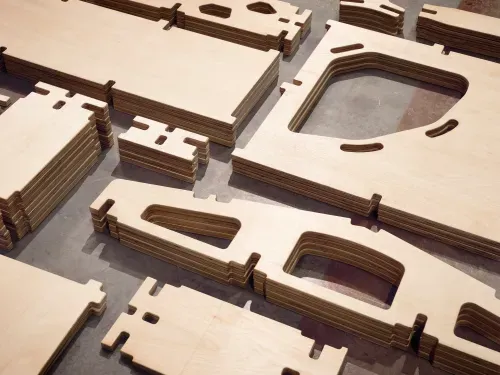 Stacked unfinished wooden cutout pieces with various shapes and slots on a concrete floor.