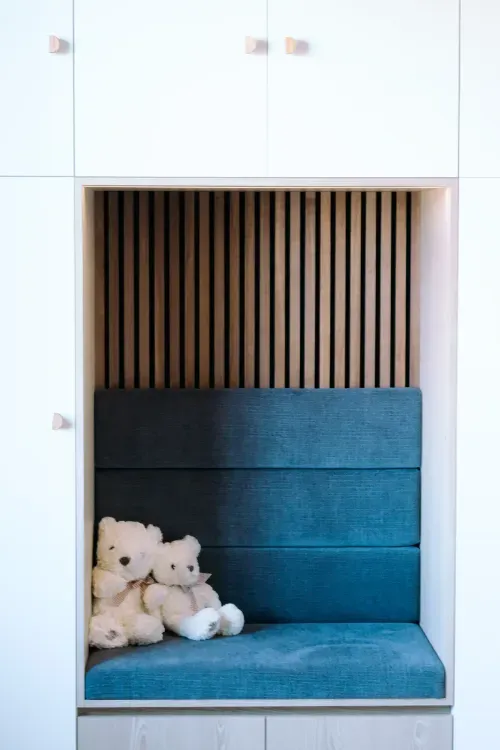Two white teddy bears sitting on a blue cushioned bench within a wooden nook with vertical slats behind.