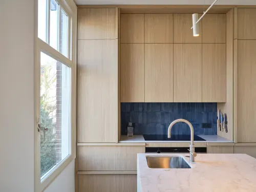 Modern kitchen with light wood cabinets, blue tiled backsplash, marble countertop island with stainless steel sink, and a large window.