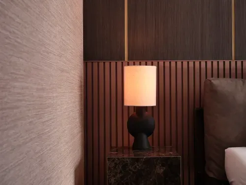 Modern bedside lamp with a black sculptural base and beige shade on a dark marble nightstand, next to a brown cushion.