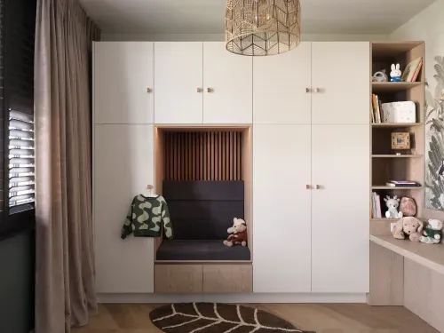 Modern built-in storage with white cabinets, a cushioned bench with a teddy bear, shelves holding toys and books, and a leafy area rug on wooden floor.