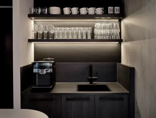 Modern kitchenette with a black coffee machine, black sink, and two shelves holding clear wine glasses, beer glasses, cups, and plates.