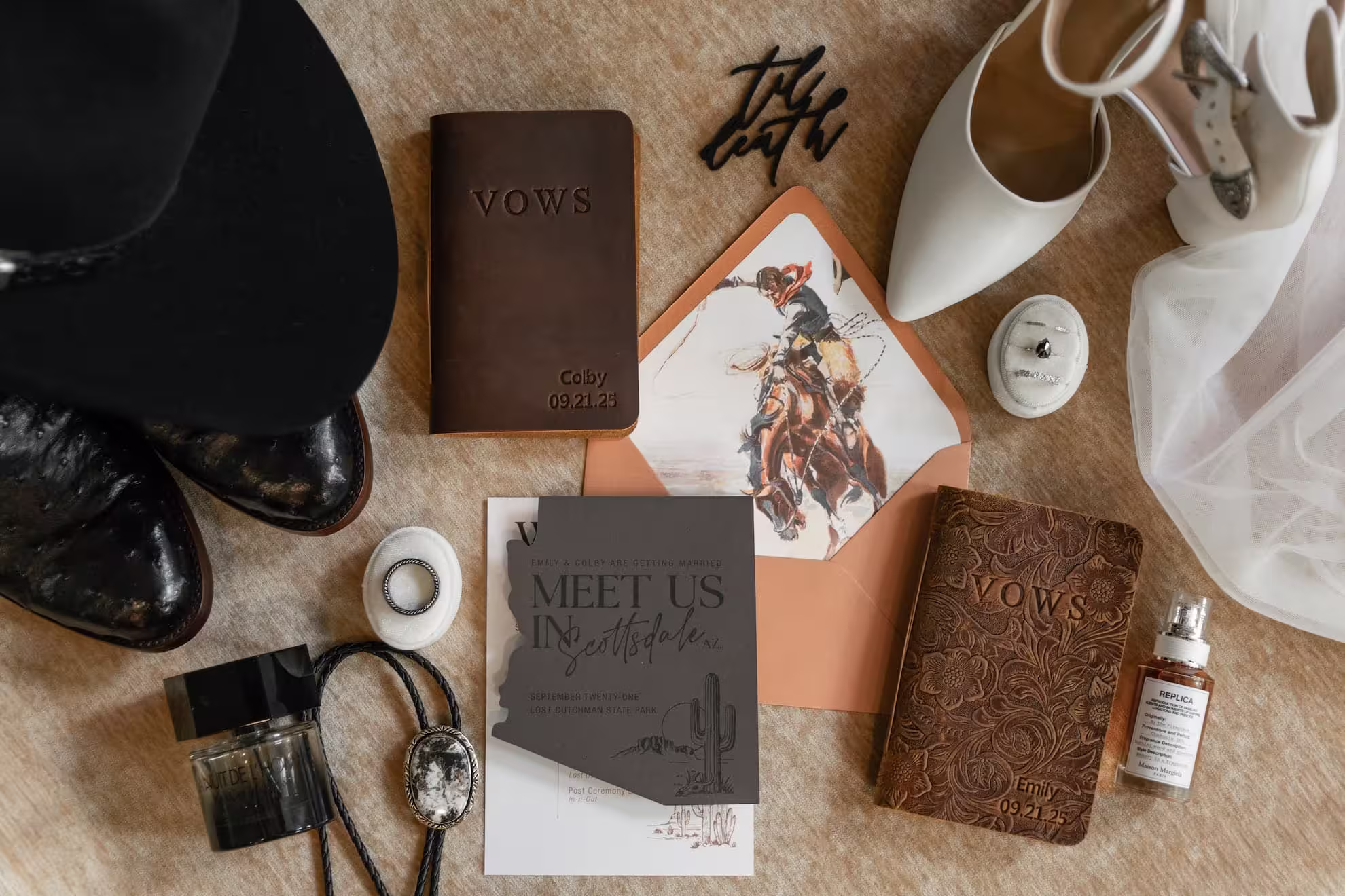 Flat lay of Arizona wedding details including custom vow books, wedding invitation suite, cowboy boots, bridal shoes, wedding rings, veil, cologne, and western-inspired accessories for a Scottsdale wedding.
