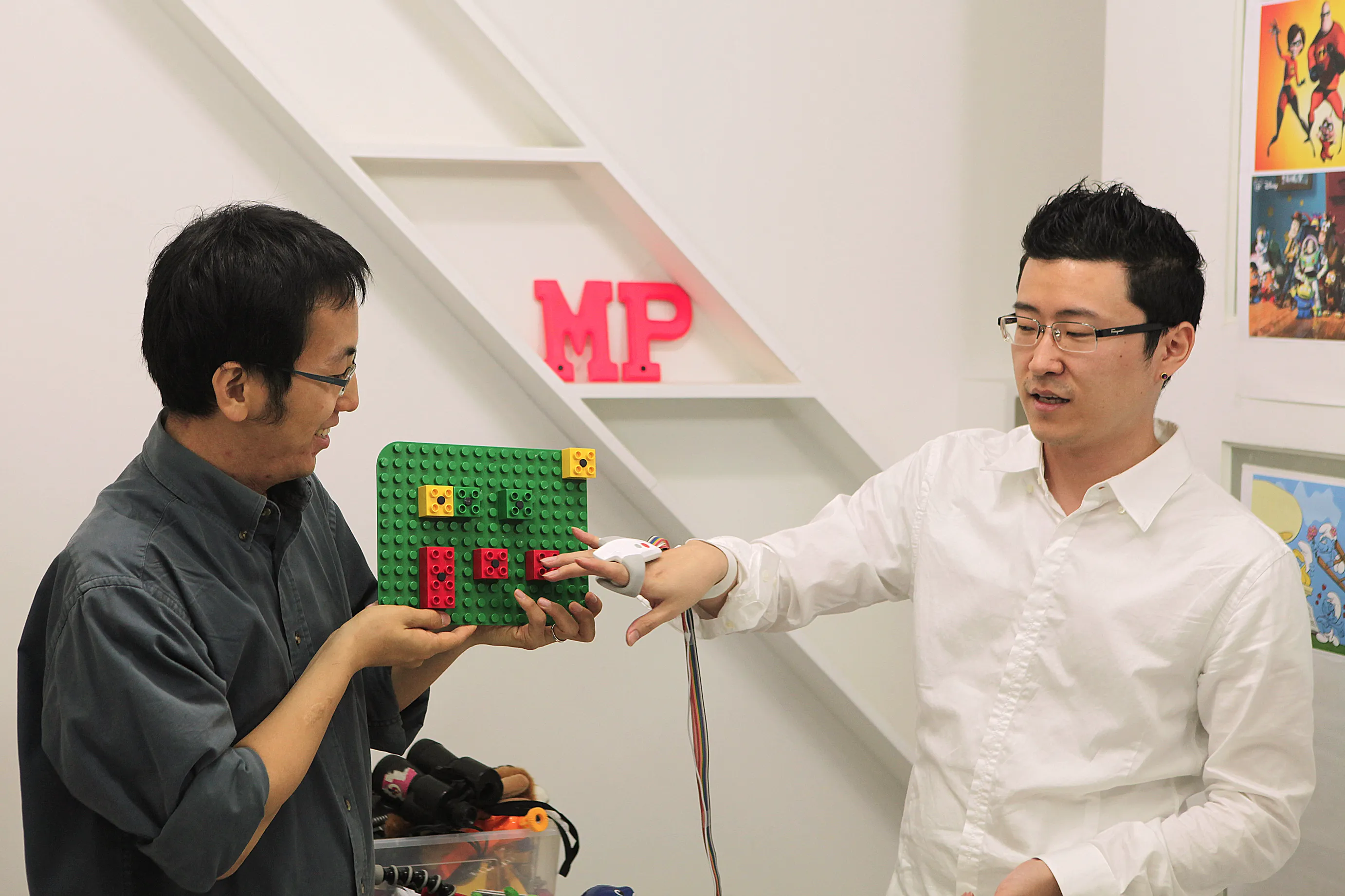 Two users demonstrate a tangible interaction prototype using a LEGO-based board; the person on the right wears an OnObject sensing device on their right hand while interacting with physical blocks.