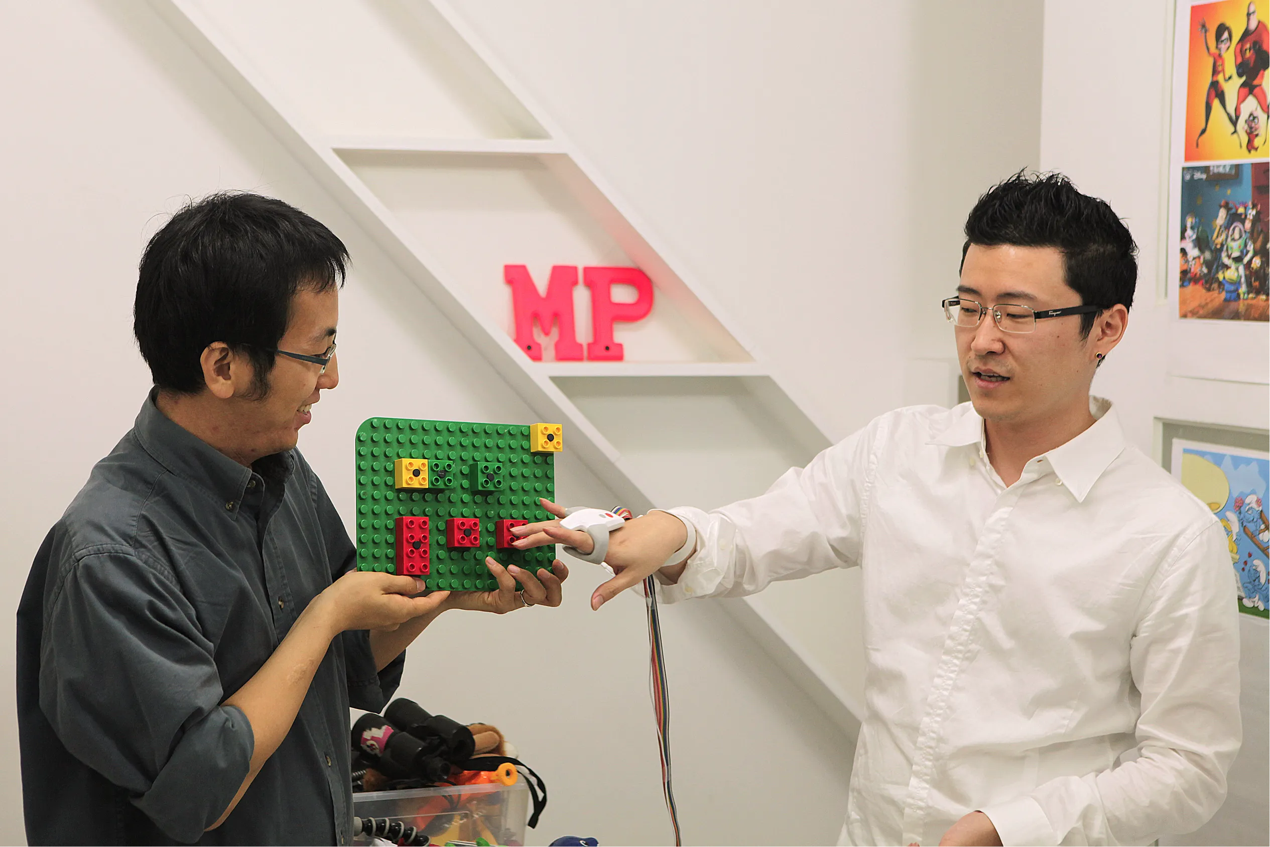 Two users demonstrate a tangible interaction prototype using a LEGO-based board; the person on the right wears an OnObject sensing device on their right hand while interacting with physical blocks.