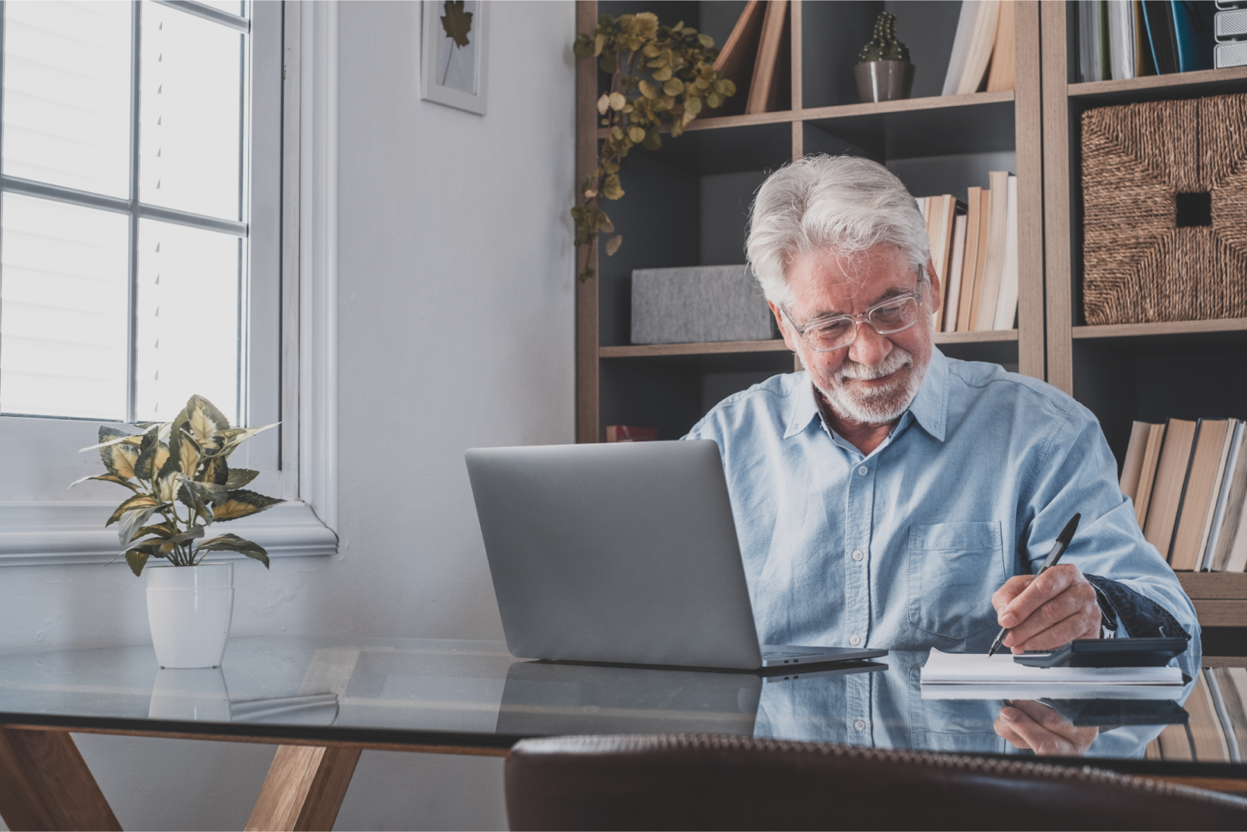 An older man smiling contently while working on his laptop and writing in his notebook.