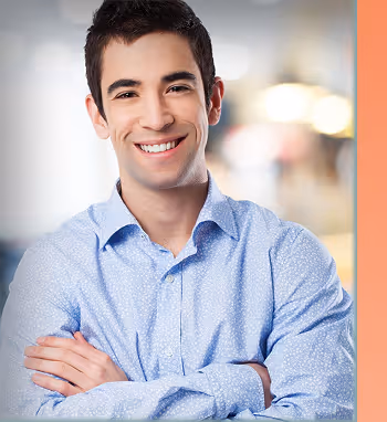 Young man with short dark hair smiling with arms crossed, wearing a light blue patterned shirt against a blurred background.