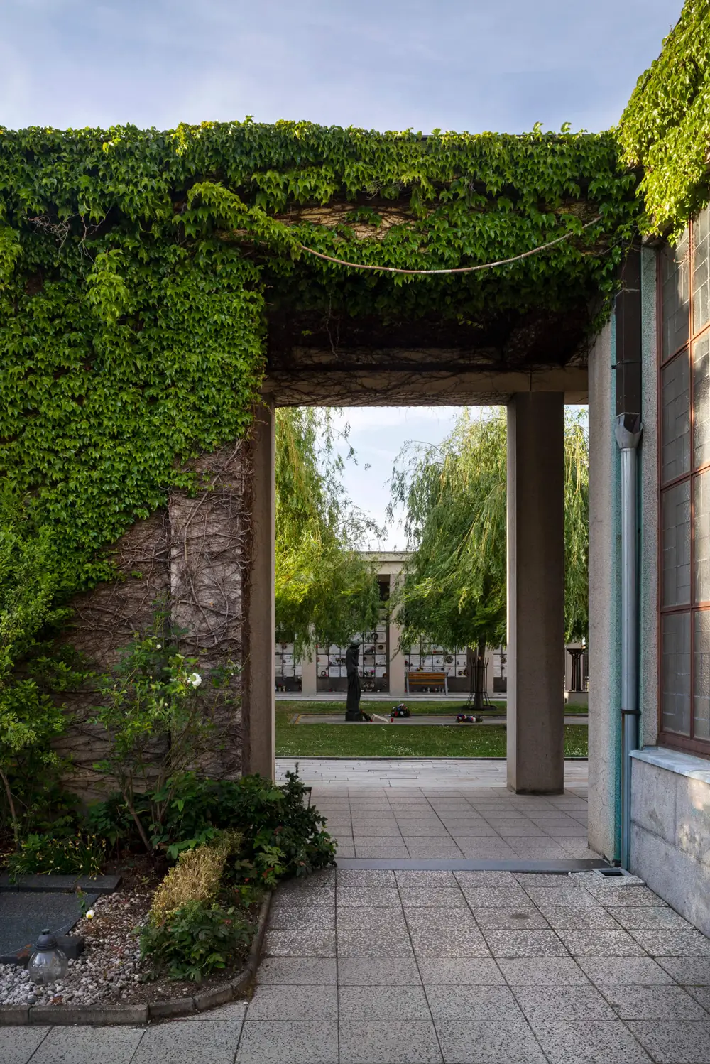 Průchod Strašnického krematoria porostlý zelení s výhledem do areálu kolumbária, architektura obklopená přírodou.