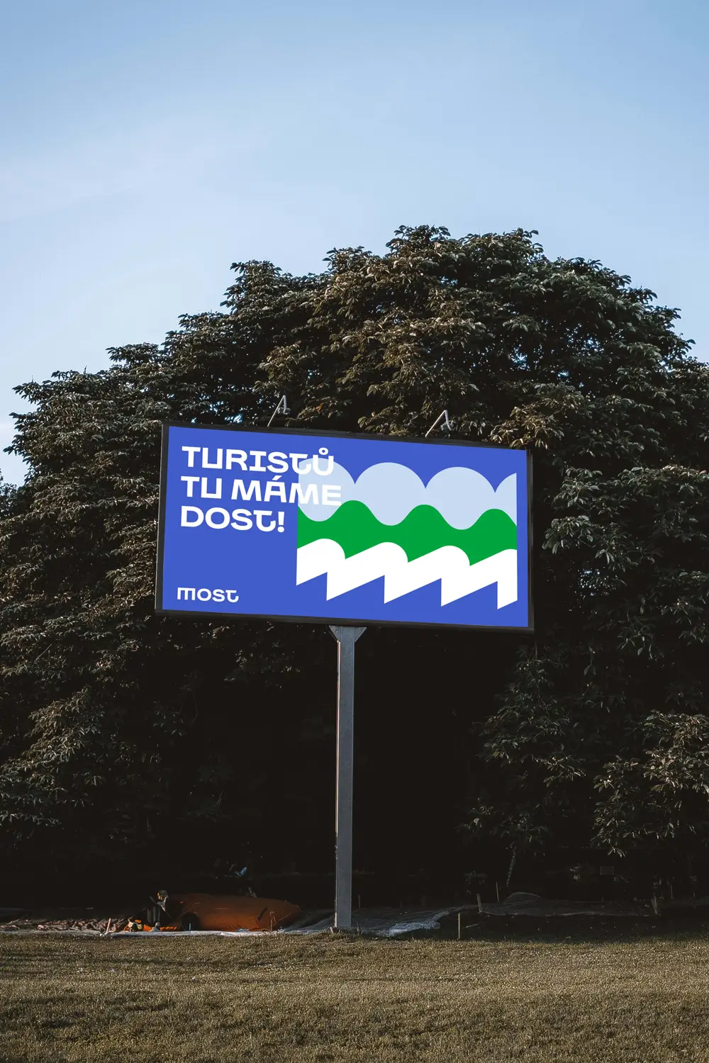 Billboard města Most s grafikou krajiny a sloganem „Turistů tu máme dost!“, instalovaný v přírodním prostředí.