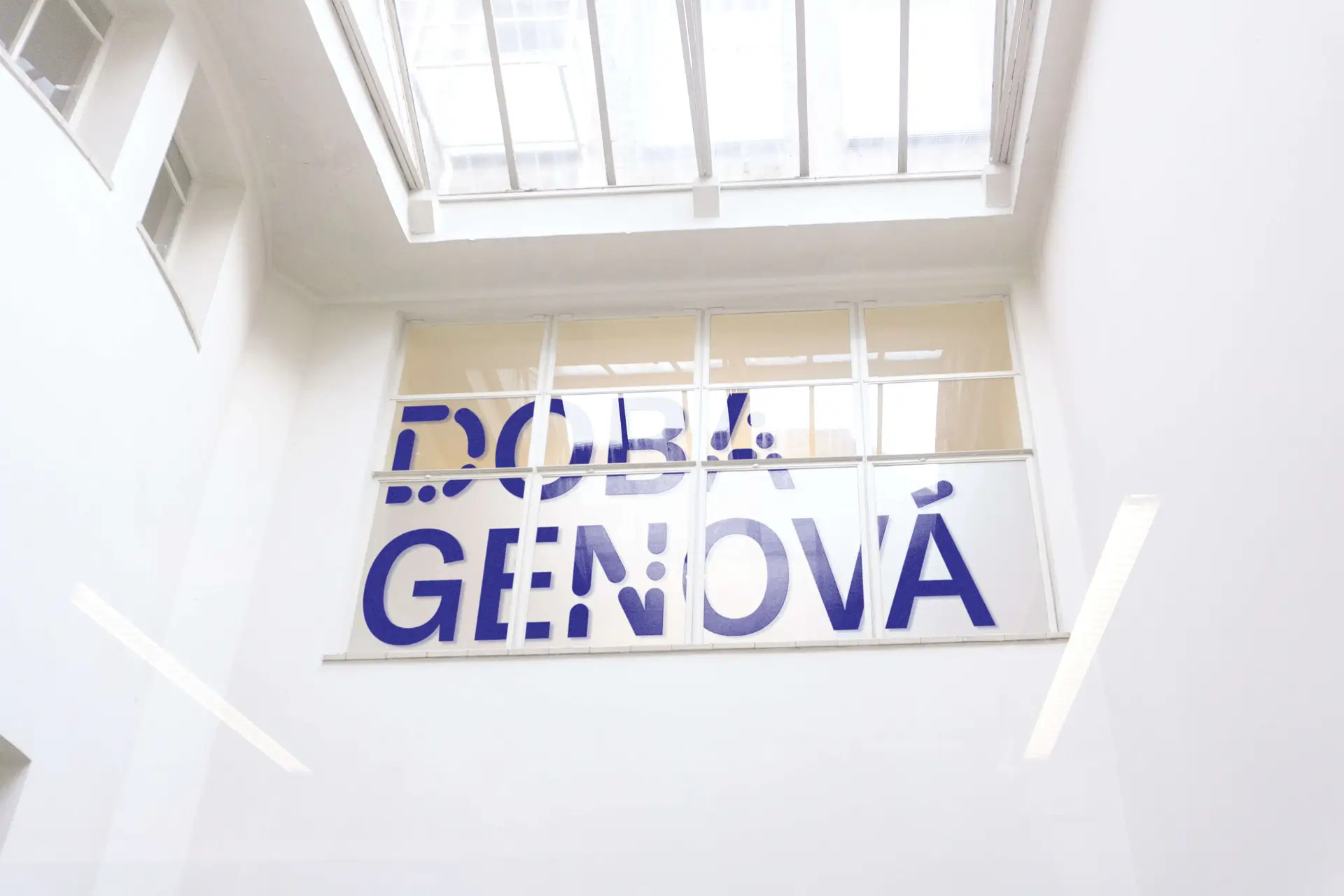 Modrý nápis Doba genová na velkoformátovém skle výstavní budovy.