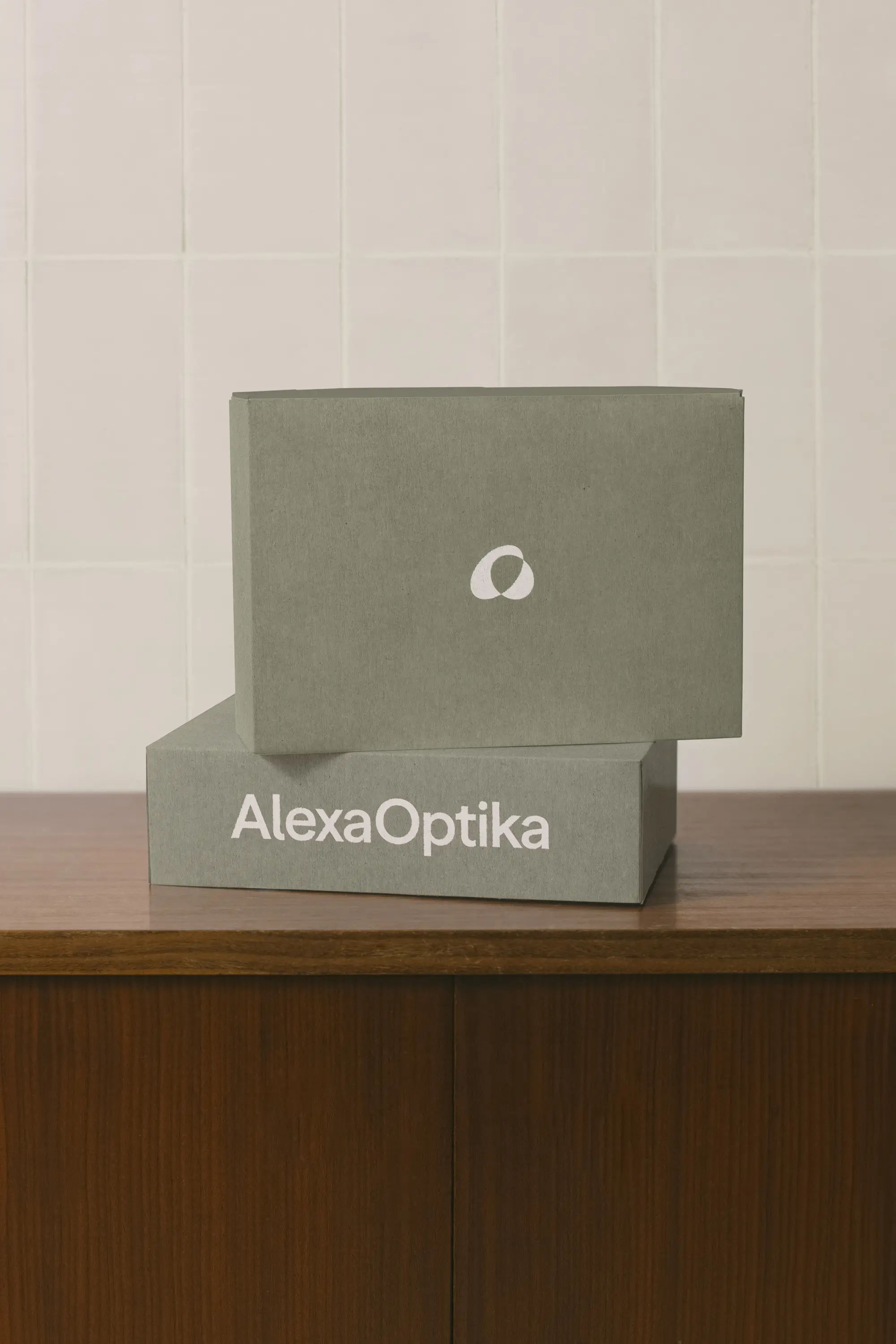 Dvě krabičky Alexa Optika v tlumeném olivovém odstínu s logem a symbolem značky, umístěné na dřevěném stole.