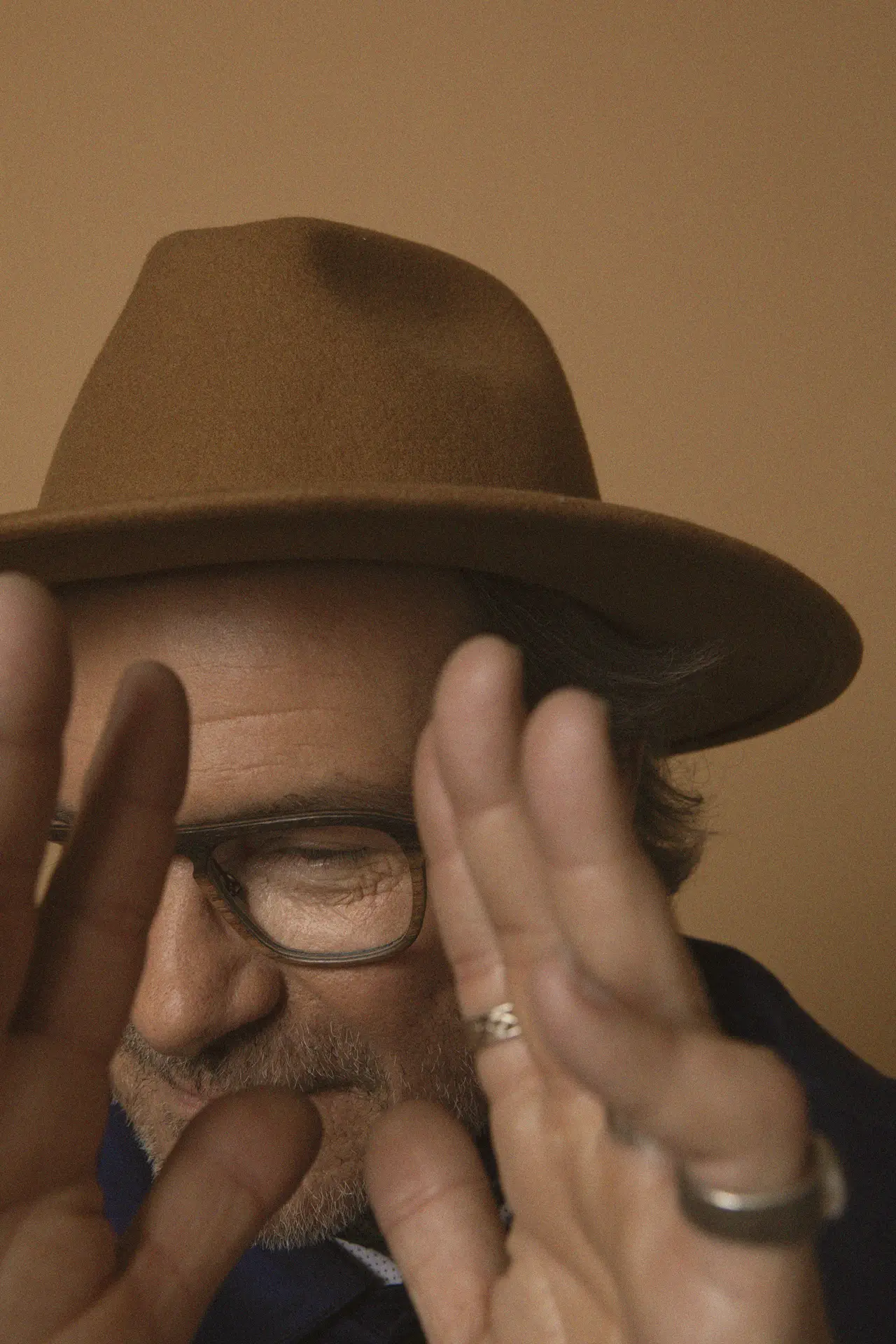 Portrét muže s brýlemi Alexa Optika a kloboukem, přirozené světlo a teplý odstín pozadí.