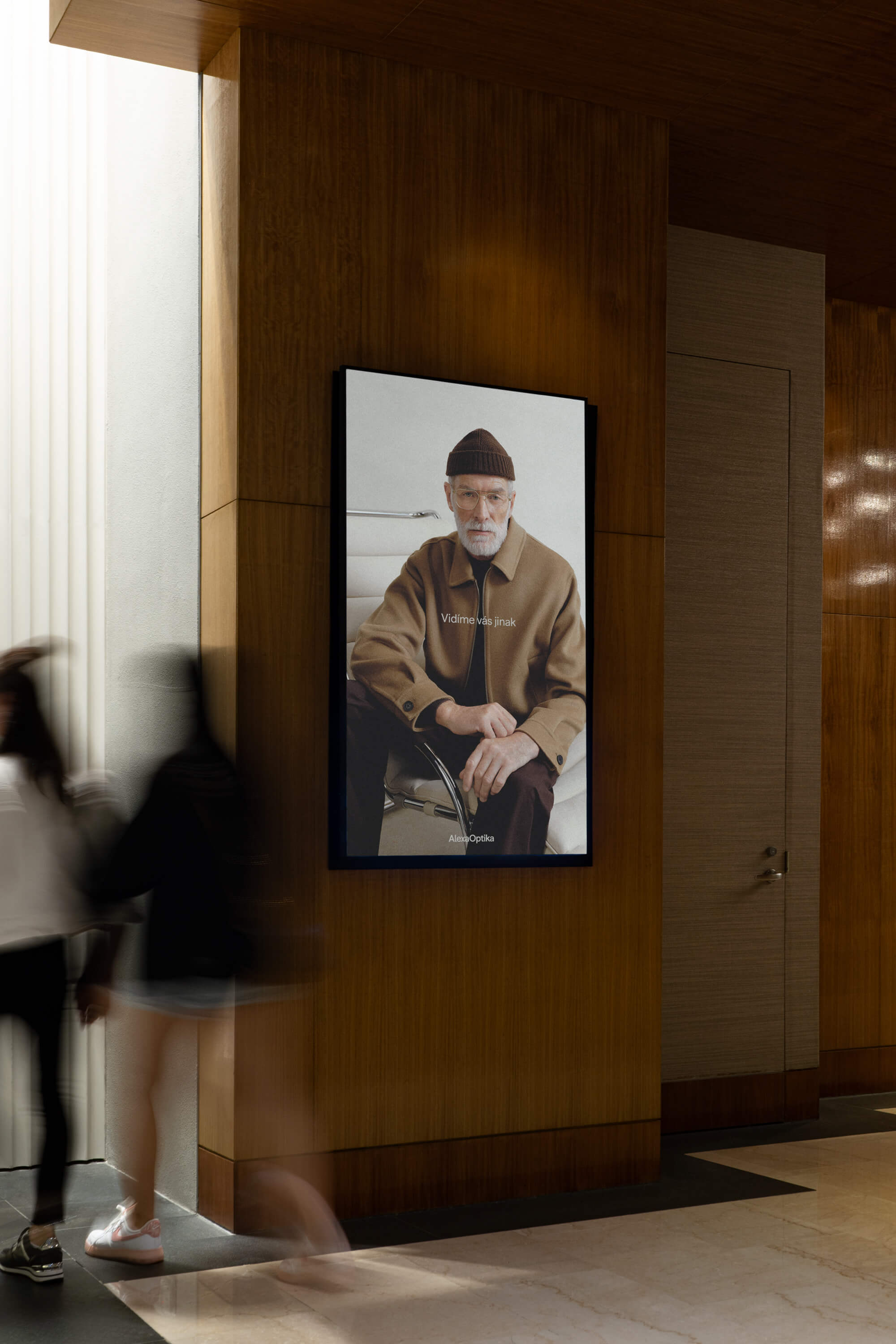 Digitální plakát kampaně Alexa Optika s portrétem staršího muže a sloganem „Vidíme vás jinak“ umístěný v interiéru s dřevěným obložením.