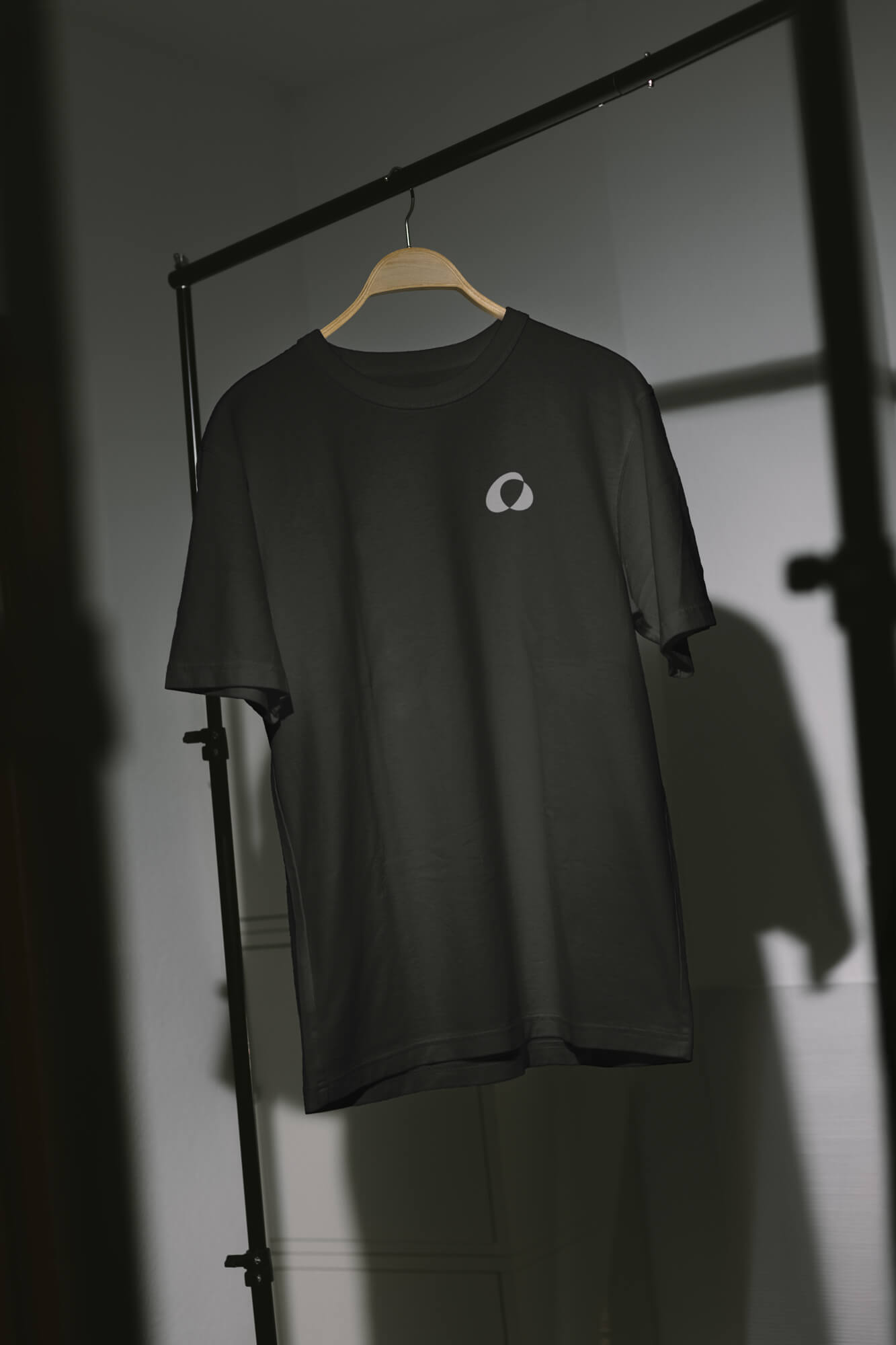 Černé tričko s logem Alexa Optika zavěšené na ramínku v jemném světle interiéru.