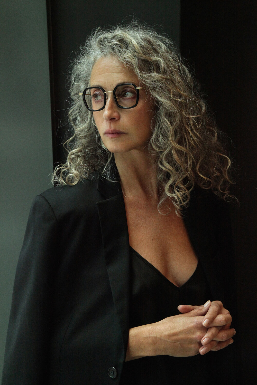 Portrét ženy s šedivými kudrnatými vlasy v brýlích z nabídky Alexa Optika, foceno v přirozeném světle.