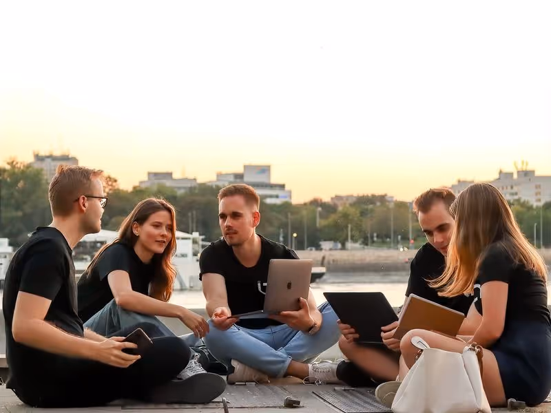 Öt fiatal ül a vízparti rakparton, laptopokkal és jegyzetfüzetekkel, naplementében beszélgetnek.