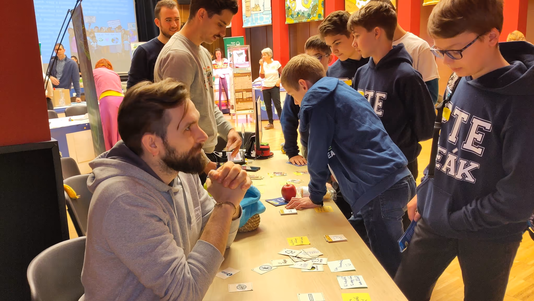 Egy férfi szürke kapucnis pulóverben ül egy asztalnál, amelyen kártyák és egy alma vannak, miközben fiatalok csoportja figyelmesen körülállja.