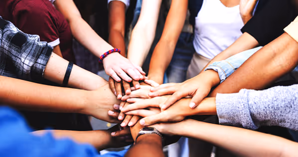 Különböző bőrszínű emberek összekulcsolják a kezüket egymáson, csapatmunkát és összetartást ábrázolva.