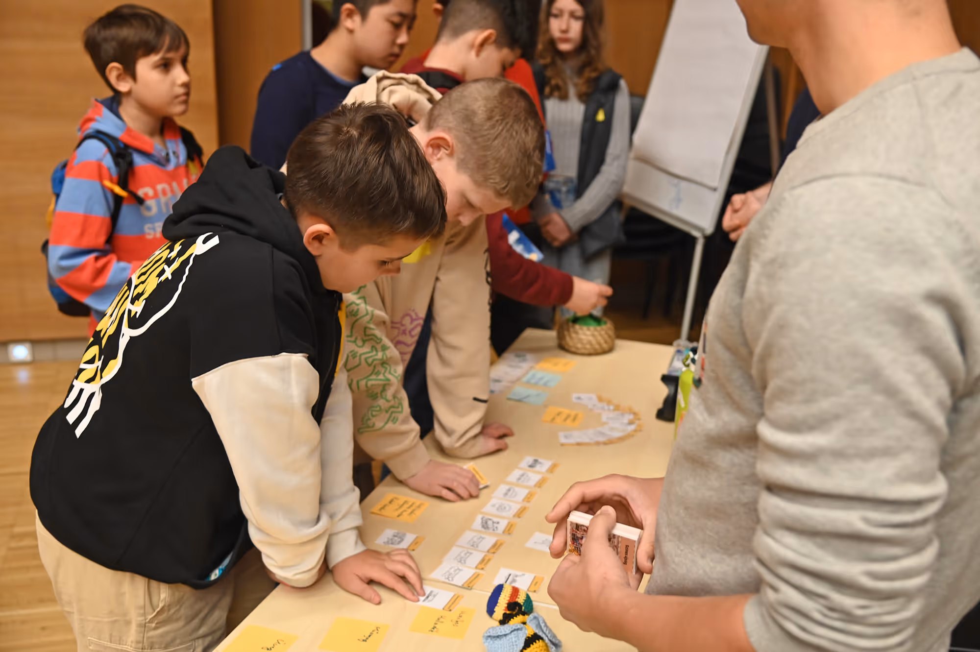 Gyerekek és egy felnőtt egy asztal körül kártyákat és jegyzeteket néznek egy beltéri oktatási foglalkozáson.