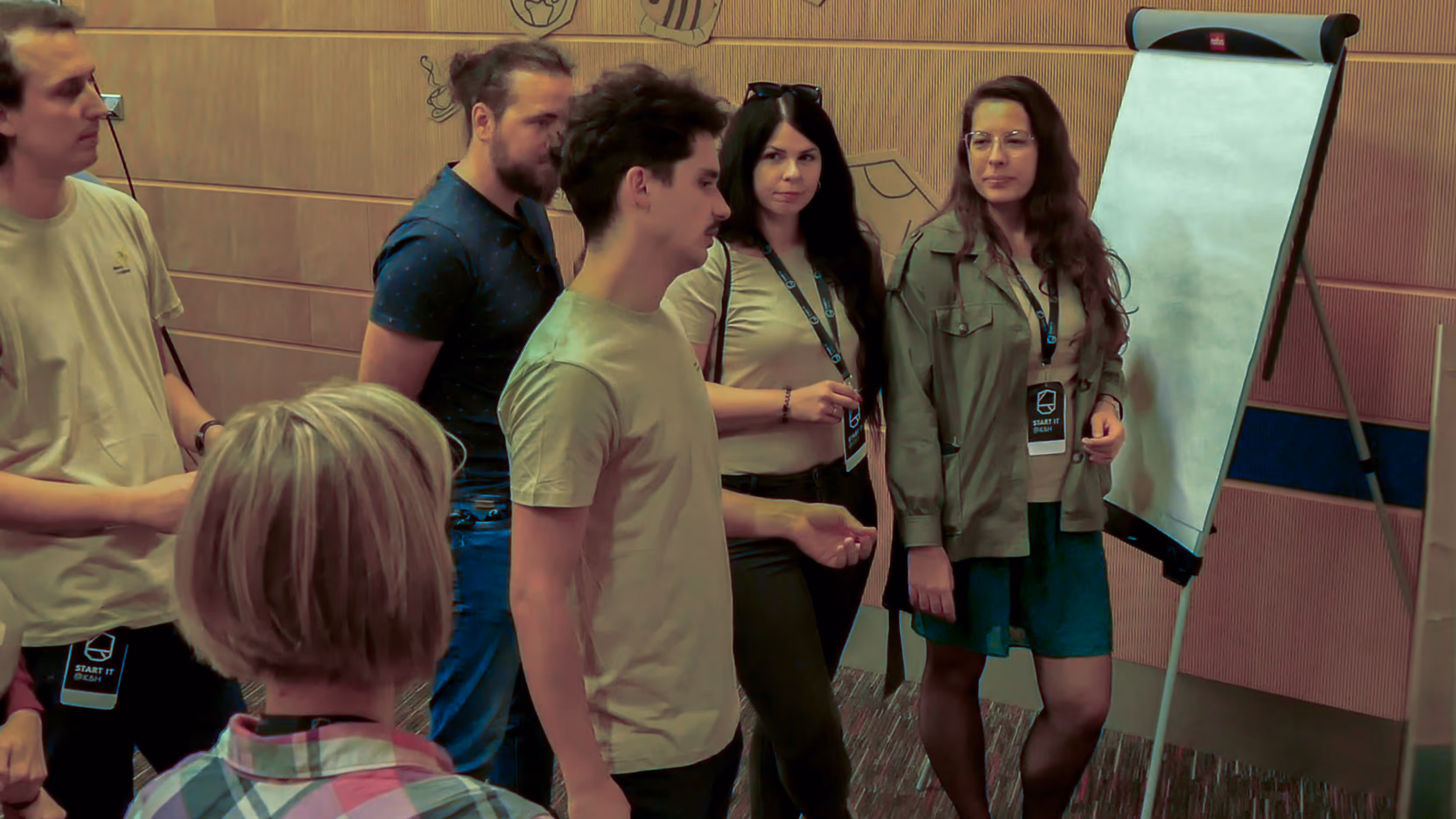 Emberek egy kis csoportja áll egy üres flipchart tábla előtt egy belső helyiségben, beszélgetés közben.