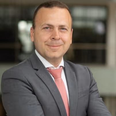 Férfi középkorú üzleti öltözetben, szürke öltönyben és rózsaszín nyakkendőben, karját keresztbe téve, enyhén mosolyogva.