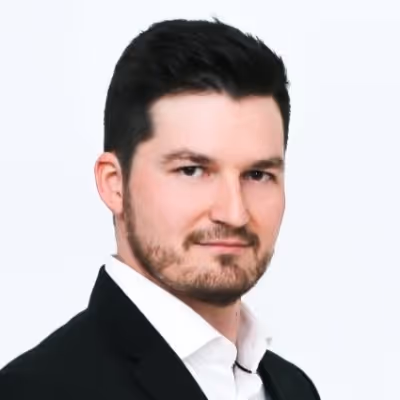 Férfi fekete öltönyben fehér inggel, rövid sötét hajjal és szakállal, világos háttér előtt.