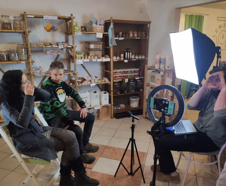 Két ember ül egy szoba egyik felében, miközben egy harmadik személy laptop és gyűrűs világítás mellett videofelvételt készít róluk.