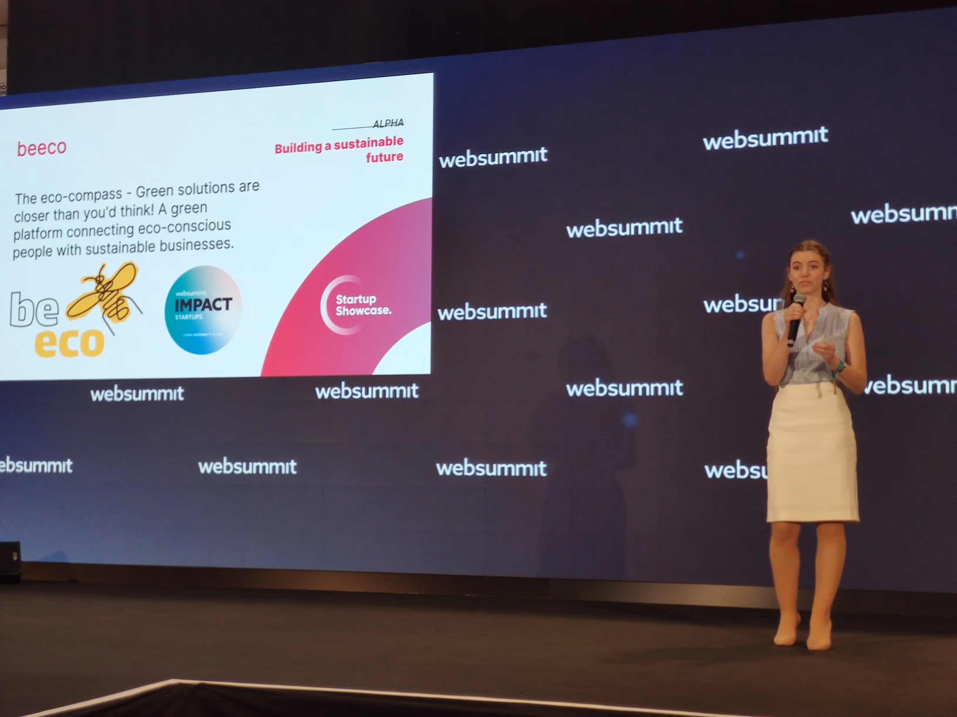 Nő áll mikrofonnal a Websummit rendezvényen, mellette egy prezentációs képernyő a beeco környezetbarát startup bemutatójával.