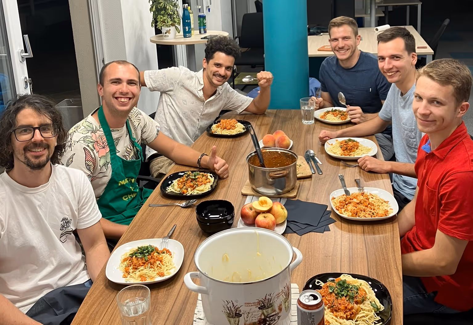 Hat mosolygó férfi ül egy asztal körül egy tál spagettit fogyasztva egy közös étkezésen, az asztalon tálak, alma és egy lábas étel van.
