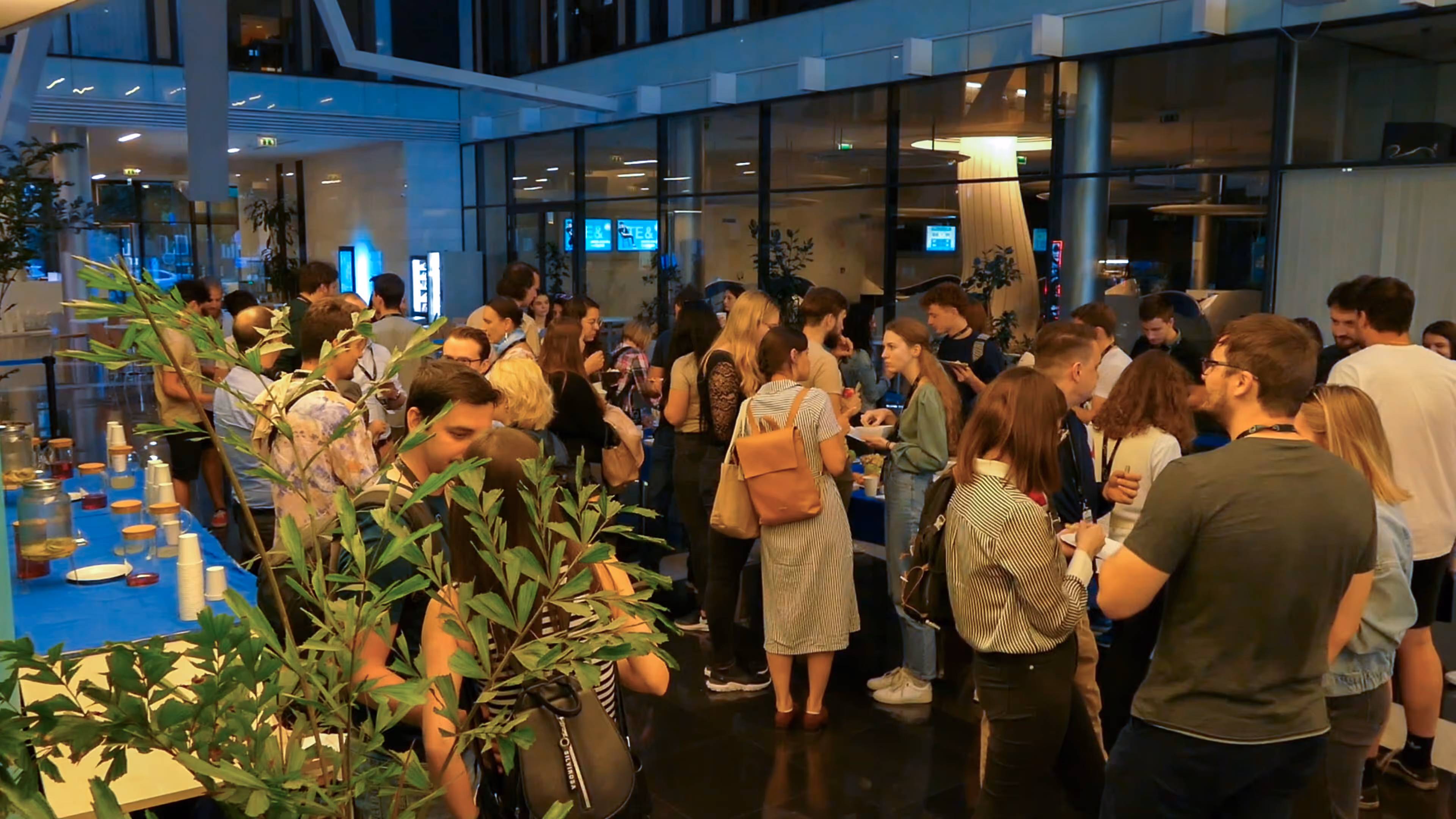 Emberek társasági eseményen egy beltéri helyszínen, asztalok italokkal és üdítőkkel.