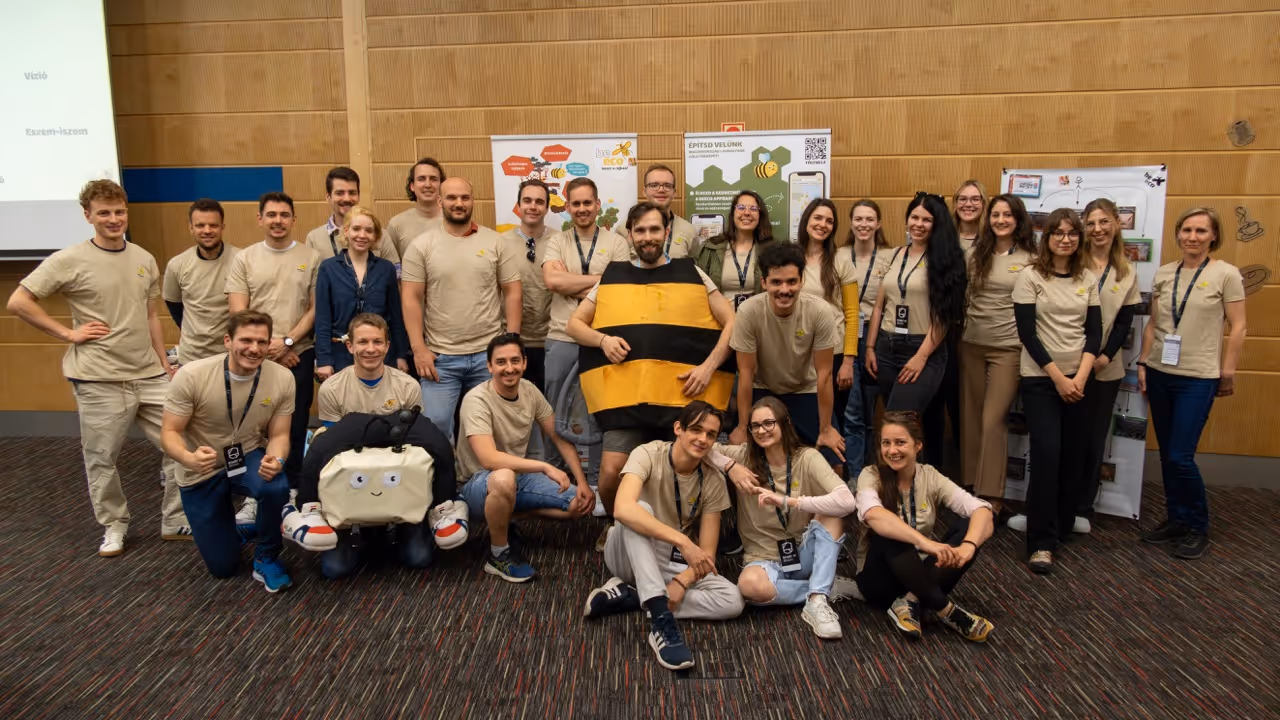 Csoportkép egy 29 fős csapatról, akik világos bézs pólót viselnek, középen egy ember méhkaptár jelmezben áll, a háttérben táblák láthatók.