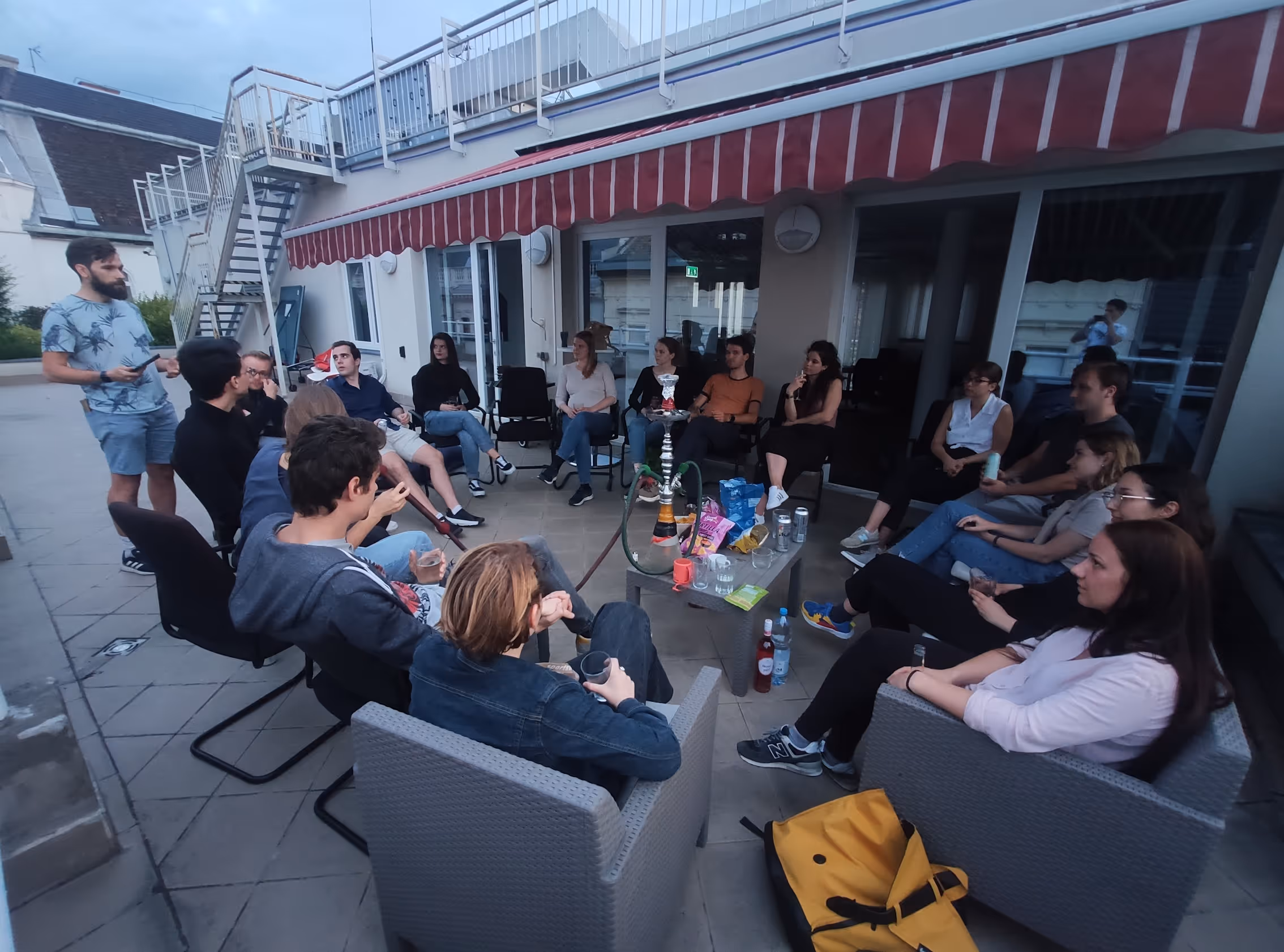 Csoport fiatal felnőttek egy tetőteraszon, akik körbe ülnek egy vízipipát és különféle italokat, chipset és nassolnivalókat tartalmazó asztalt.