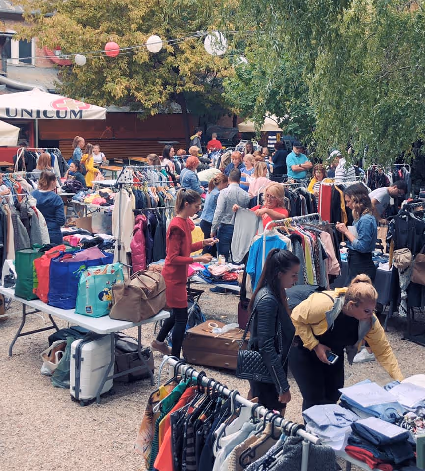 Emberek egy szabadtéri bolhapiacon ruhákat nézegetnek és válogatnak árusító asztalok és ruhássállványok között faágak alatt.