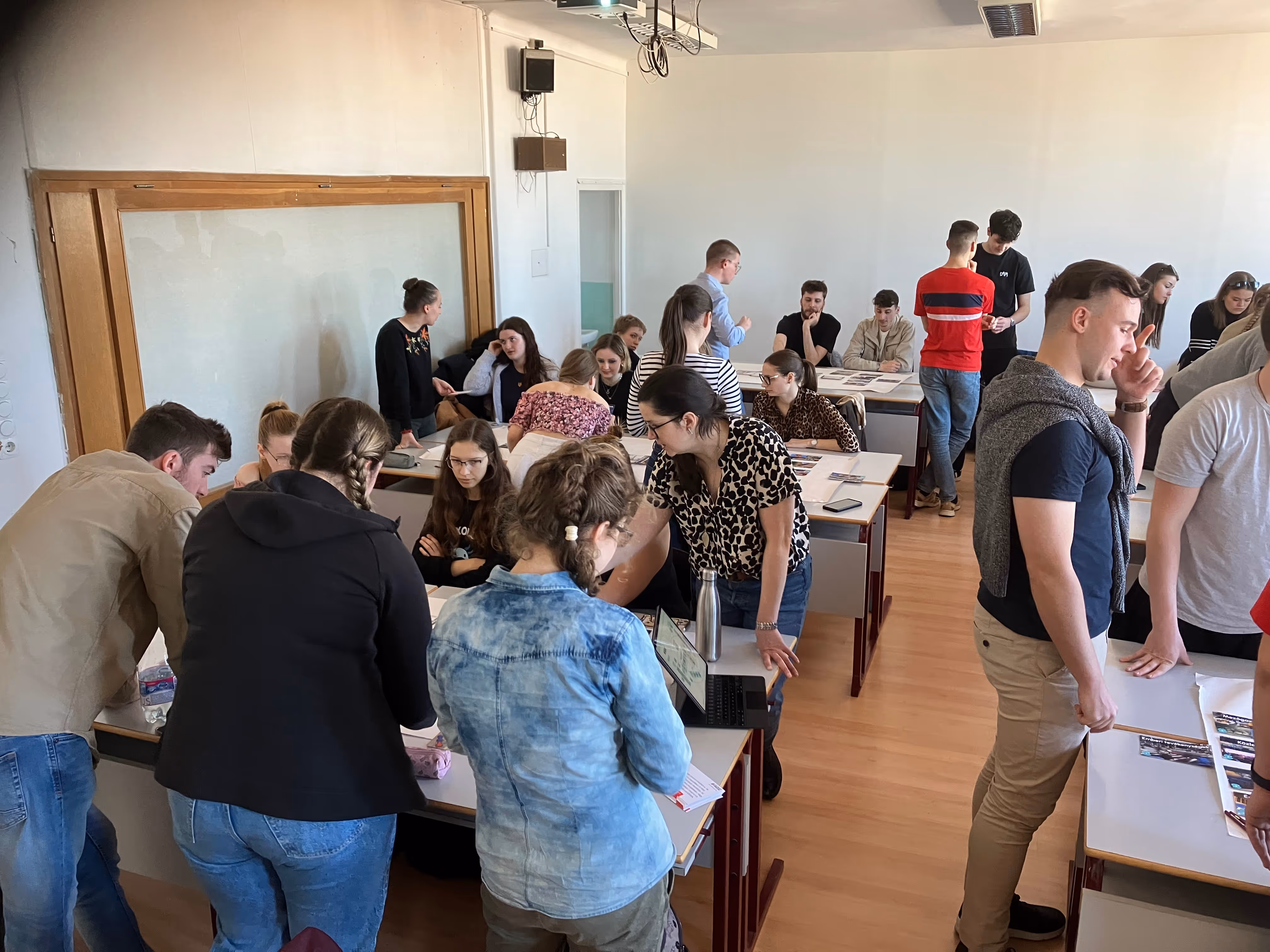 Diákok csoportjai egy osztályteremben különböző asztaloknál beszélgetnek és dolgoznak együtt napsütéses napon.