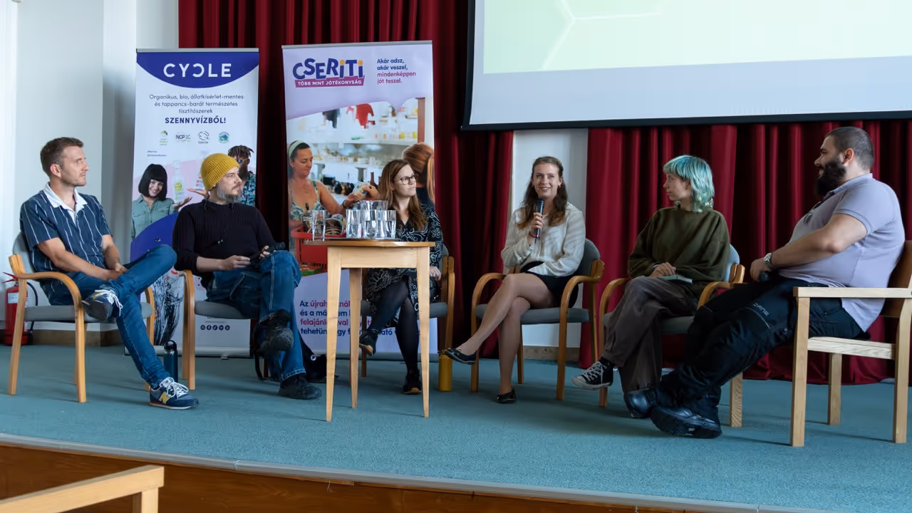 Hat ember ül egy színpadi beszélgetésen, egyik nő mikrofont tart, háttérben két információs molinó és piros függöny.