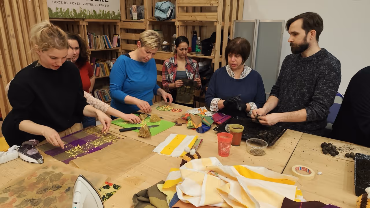 Emberek egy asztal körül, különböző kézműves anyagokkal dolgoznak, köztük textil, földlabdák és vasalók.